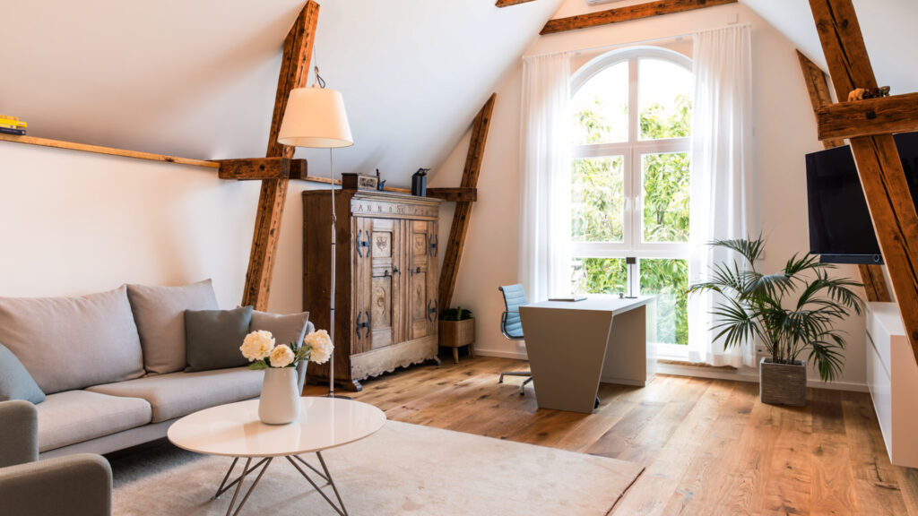 Helles, modern eingerichtetes Wohnzimmer mit sichtbaren Holzbalken, einem historischen Holzschrank, einem schlichten Schreibtisch am Fenster und gemütlicher Sofaecke mit Couchtisch und Blumen.